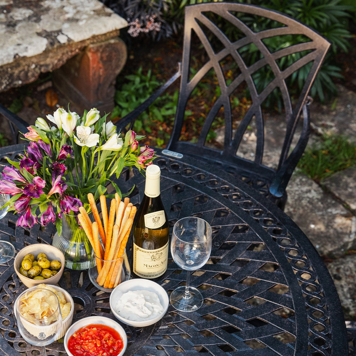 Outdoor round garden table and chair with a bottle of wine, glasses, appetizers, and flowers on a flagstone patio.