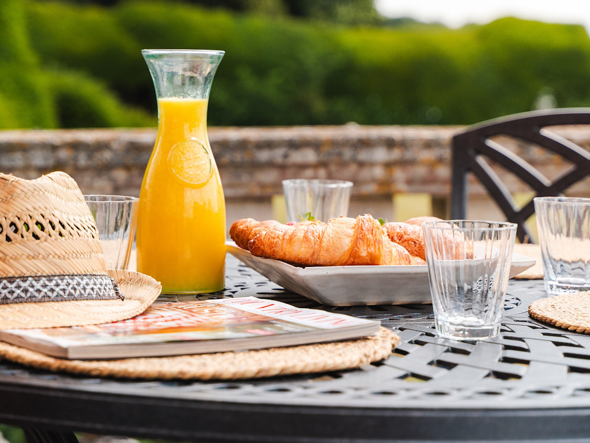 Croissants, Orangensaft, Trinkbecher und Strohhut mit Zeitschrift auf Gartentisch